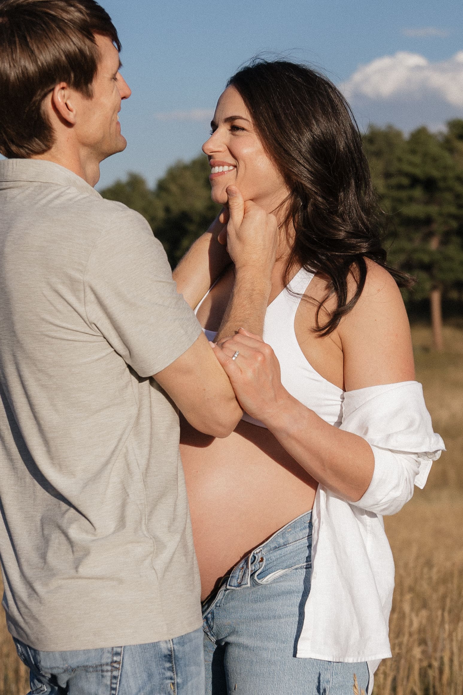 Maternity session at Betasso Park featuring an expecting couple in golden grass with rolling hills, pine trees, and blue sky with clouds, ending with sunset portraits overlooking Boulder.
